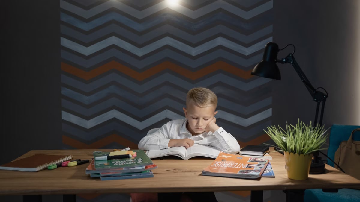 Young boy focused on reading at a desk filled with books and a lamp in dim lighting.